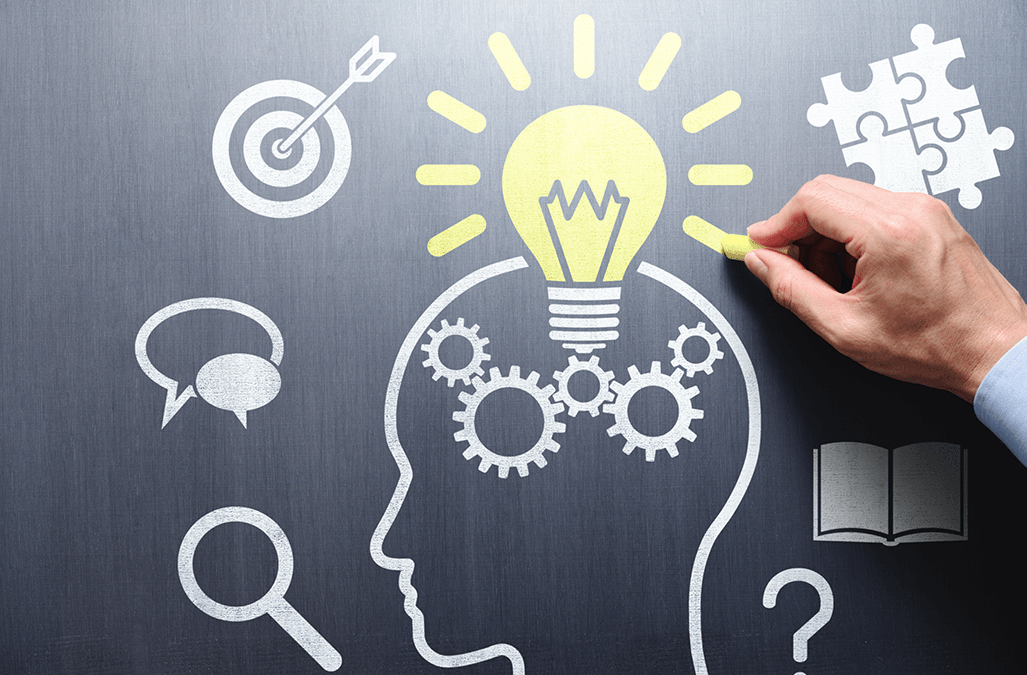 Photo of a hand drawing on a chalkboard, image is the outline of a head with cogs inside surrounded by puzzle pieces, speech bubbles, magnifying glass and a book, with a lightbulb from the top of the head.
