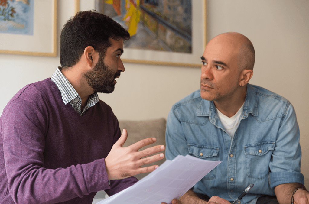 Photo of man with papers explaining to man beside him on a couch
