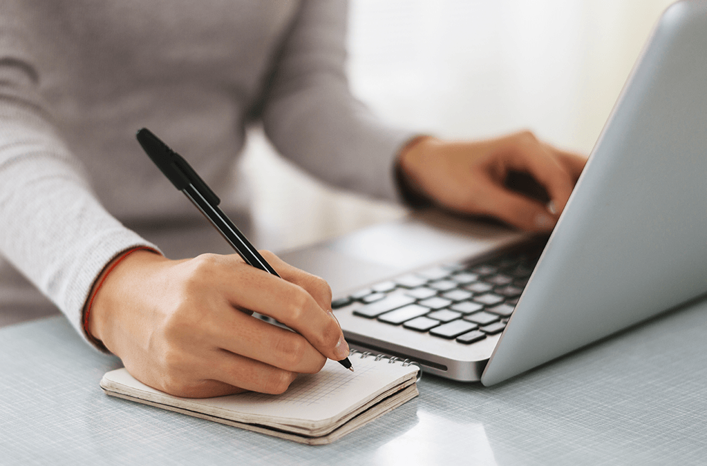Photo of a woman at laptop with pen to small notepad