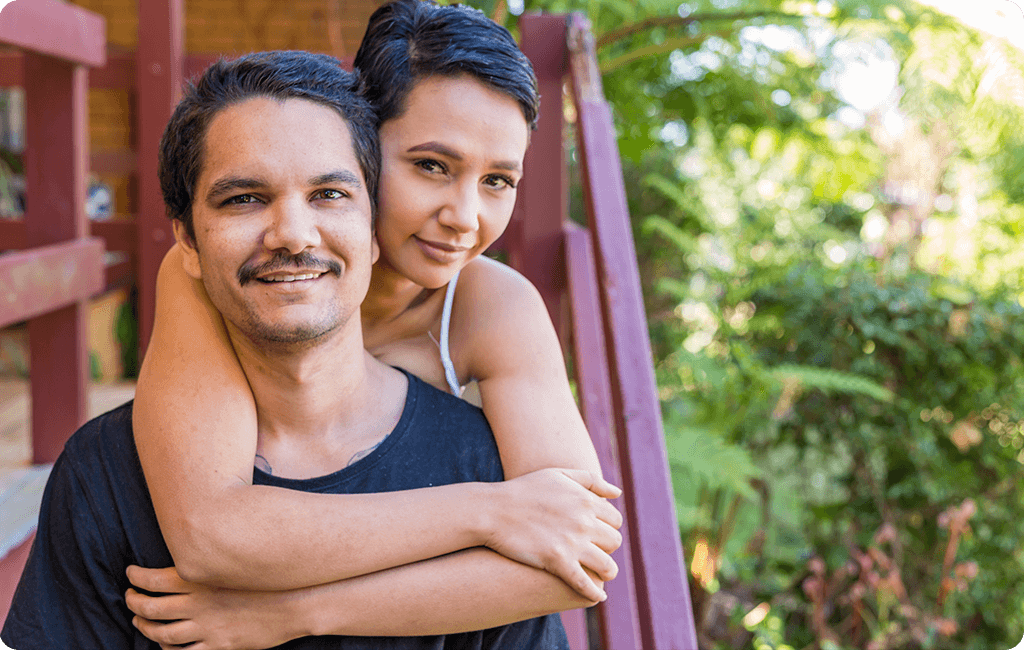 Image of young Indigenous couple on back stairs of a house with green ferns behind them.