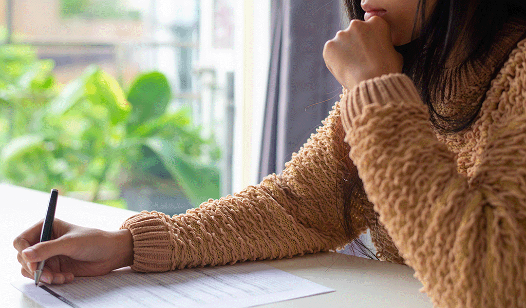 Woman in mustard coloured jumper filling in a form at a table with plants in background.