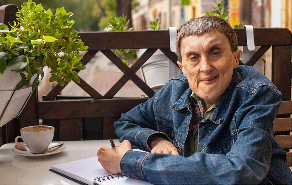 A middle-aged person with genetic disability sitting at a table with a cup of coffee and a notebook